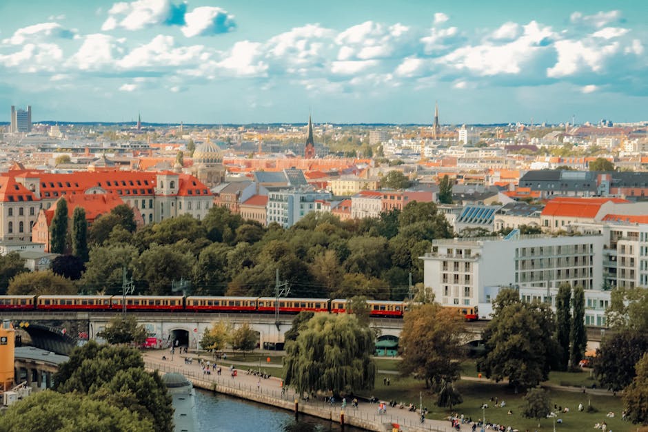 A picturesque cityscape of Berlin featuring a train, lush greenery, and vibrant architecture under a bright sky.